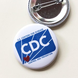 May include: A white button with the text "U.S. CENTERS FOR DISEASE CONTROL AND PREVENTION" and the letters "CDC" in blue. A small red heart is visible. The back of the button is silver with a pin.