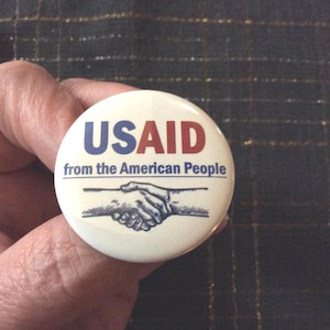 May include: A round white pinback button featuring the blue and red text USAID, the blue text from the American People, and a black and white graphic illustration of two hands shaking, centered on the button face against a plain white background.