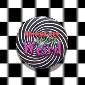 May include: A circular pin-back button featuring a black and white spiral pattern. The text "Things are" is printed in orange, "getting" in green, and "Weird" in pink with a jagged, stylized font, centered over the hypnotic spiral design.