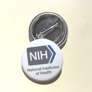 May include: A white button with the text "NIH" in gray and a blue arrow, with the words "National Institutes of Health" below. The button has a metal pin back.