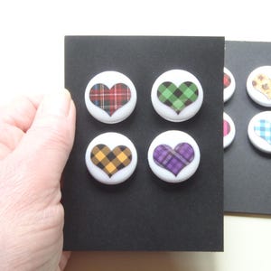 May include: A collection of heart-shaped magnets with plaid patterns in red, green, yellow, and purple. The magnets are white with a black backing. Additional magnets with floral and gingham patterns are also visible.