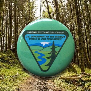 May include: A round, green button featuring the National System of Public Lands logo. The logo includes a triangle with a mountain and river scene, and the text "U.S. Department of the Interior Bureau of Land Management."