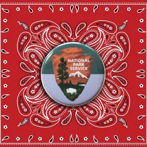 May include: A round National Park Service button featuring a mountain scene with a tree and a bison, set against a green and white background. The button is placed on a red bandana with a white paisley pattern.