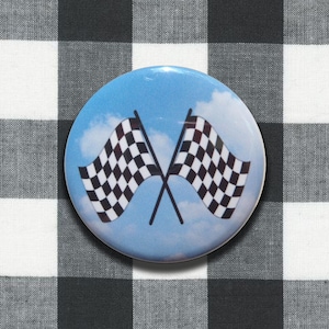 May include: A circular pin-back button featuring a graphic of two crossed black and white checkered racing flags set against a light blue sky with soft white clouds. The design is centered on the round surface of the button.