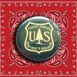 May include: A circular pinback button featuring a dark green background with the U.S. Forest Service shield logo in cream. The logo displays a pine tree between the letters U and S, with the text Forest Service Department of Agriculture.