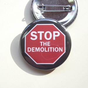 May include: A round pin-back button featuring a red octagonal stop sign graphic with a white border and the white text STOP THE DEMOLITION centered on the front. The back of the button is metallic silver with a horizontal pin fastener attached.