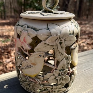 May include: A ceramic jar with a lid, featuring a relief design of birds and flowers. The jar is off-white with green and pink accents. The lid has a loop handle. The jar has cut-out sections.