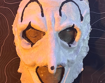 May include: A white, textured mask with black outlines and eye sockets. The mask has a clown-like appearance with a wide, open mouth and a small, round nose. The mask is likely handmade and has a slightly distressed look.