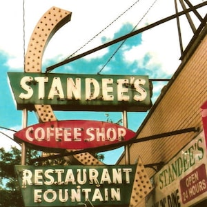 May include: A vintage neon sign for "Standee's" coffee shop, restaurant, and fountain. The sign is green, red, and white with a curved shape and the text "Standee's Coffee Shop Restaurant Fountain" in white lettering.