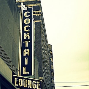 May include: A vintage sign with black and white lettering that reads "Cocktail Lounge". The sign is mounted on a brick building with a faded, vintage look.