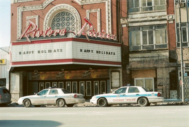 May include: A white police car with the words "Chicago Police" on the side is parked in front of a building with a sign that reads "Riviera" and "Happy Holidays".