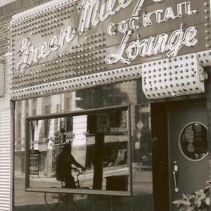 May include: A sepia-toned photograph of the Green Mill Cocktail Lounge storefront. The building features a large neon sign with the text Green Mill Cocktail Lounge, a grid of light bulbs, a large glass window reflecting a cyclist, and a door.