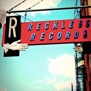 May include: A rectangular hanging sign for Reckless Records featuring a red background with blue lettering. A large white letter R is on the left side, accompanied by a white sculpted hand pointing outward. A second white hand is visible on the right side.