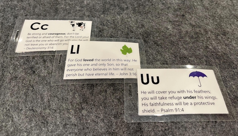 Może przedstawiać: Trzy kolorowe karty z nadrukowanymi literami "Cc", "Li" i "Uu". Każda karta ma inny obrazek i wydrukowany werset biblijny. Karta "Cc" ma krowę, karta "Li" ma zielony liść, a karta "Uu" ma niebieski parasol.