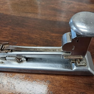 May include: A vintage silver stapler with a large, rounded top and a lever. The stapler is made of metal and sits on a wooden surface. The design is classic and functional, suitable for office or home use.