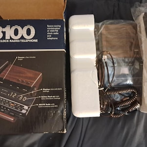 May include: A vintage GTE 8100 AM/FM clock radio/telephone in its original box. The box is dark blue with white text and a picture of the product. The product has a wood-grain finish and a digital clock display.