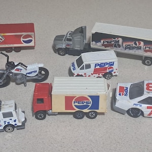 May include: A collection of Pepsi-branded toy vehicles, including trucks, a motorcycle, a van, and a race car. The vehicles are white, red, and blue, with the Pepsi logo prominently displayed on each. The race car has the number 73 on the side.