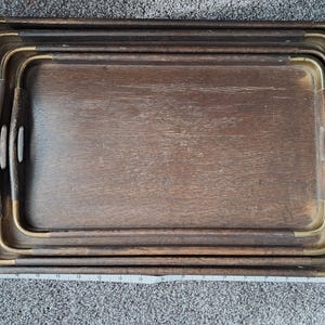 May include: Set of four wooden serving trays. The trays are nested, with a dark brown finish and brass-colored metal corner accents. Each tray has a rectangular shape with rounded corners and integrated handles.