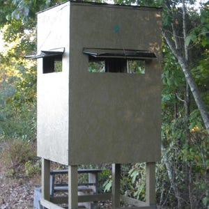 May include: A wooden hunting blind with a gray exterior and two small windows. The blind is elevated on a wooden platform with a ladder leading up to the platform.