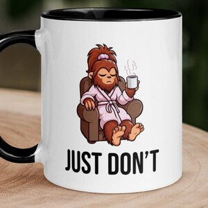 May include: A white ceramic mug with a black handle and rim featuring a graphic of a brown creature with a ponytail wearing a pink robe, sitting in a brown armchair while holding a steaming mug, with the black text JUST DON'T printed below.