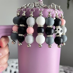 May include: A collection of beaded cup charms in various colors, including gray, black, white, and pink, attached to a silver chain. The charms are displayed on a lavender tumbler with a matching straw.