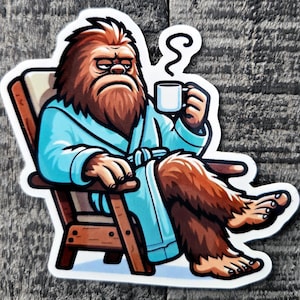 May include: A sticker featuring a cartoon Bigfoot in a blue robe, seated in a wooden chair, holding a mug of coffee. The Bigfoot has brown fur and a grumpy expression. The sticker is on a gray wooden surface.