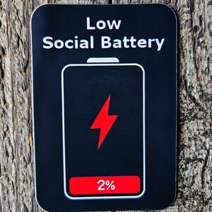 May include: A black sticker with the text "Low Social Battery" above a phone battery icon. The battery is at 2% with a red lightning bolt in the middle. The sticker is on a weathered wooden surface.