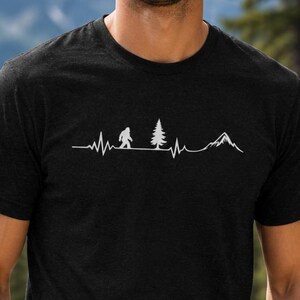 May include: A black short-sleeve t-shirt featuring a white graphic print of a heartbeat line that incorporates a silhouette of a Bigfoot, a pine tree, and a mountain peak across the chest area of the garment.