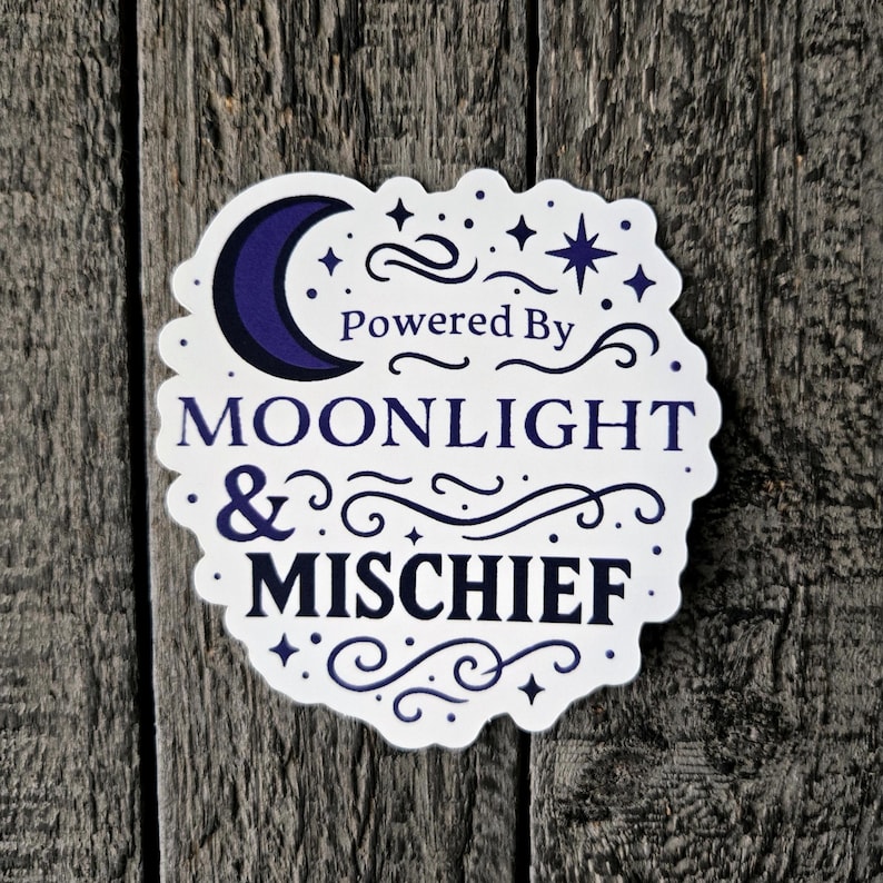 May include: A white sticker with a dark blue crescent moon, stars, and the words "Powered By Moonlight & Mischief" in a decorative font. The sticker has a scalloped edge and is set against a weathered wood background.
