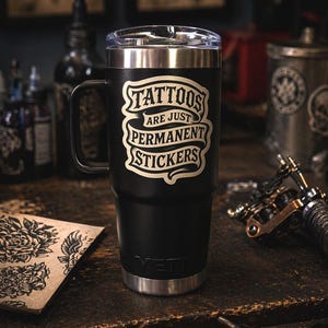 May include: A black stainless steel tumbler with a handle and a clear lid. The front features a cream-colored graphic design with the text "TATTOOS ARE JUST PERMANENT STICKERS" in a stylized banner font. The brand name "YETI" is embossed at the base.