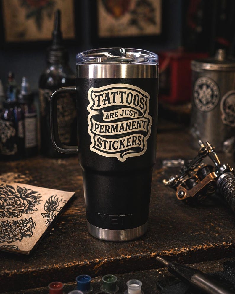 May include: A black stainless steel tumbler with a handle and a clear lid. The front features a cream-colored graphic design with the text "TATTOOS ARE JUST PERMANENT STICKERS" in a stylized banner font. The brand name "YETI" is embossed at the base.