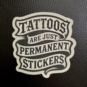 May include: A die-cut sticker featuring a black and off-white banner design with the text TATTOOS ARE JUST PERMANENT STICKERS in a bold, stylized font. The graphic has a layered appearance with shading that creates a sense of depth and dimension.