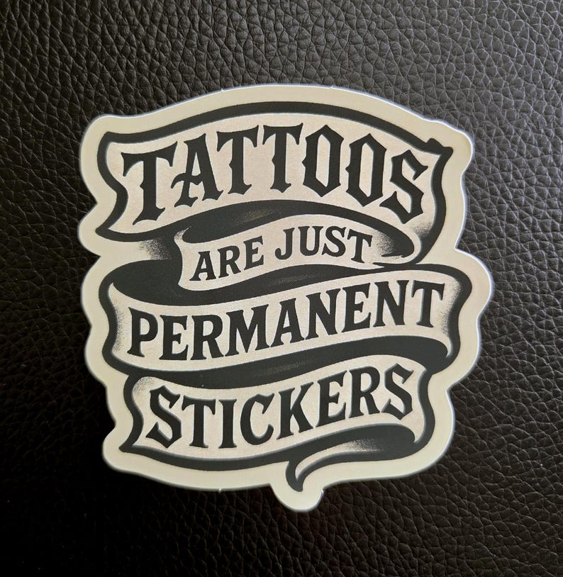 May include: A die-cut sticker featuring a black and off-white banner design with the text TATTOOS ARE JUST PERMANENT STICKERS in a bold, stylized font. The graphic has a layered appearance with shading that creates a sense of depth and dimension.