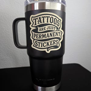 May include: A matte black stainless steel tumbler with a handle and a clear lid. The front features a cream-colored graphic design with the text "TATTOOS ARE JUST PERMANENT STICKERS" arranged in a banner style. The YETI logo is embossed at the base.