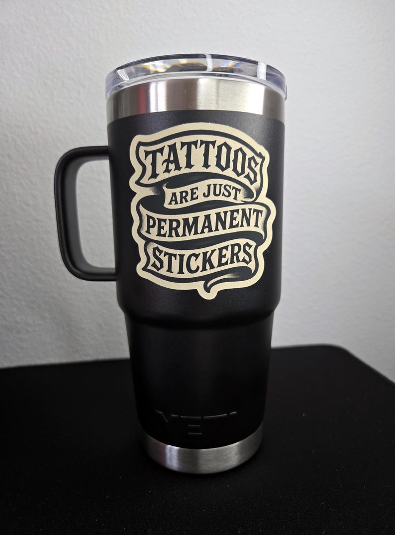 May include: A matte black stainless steel tumbler with a handle and a clear lid. The front features a cream-colored graphic design with the text "TATTOOS ARE JUST PERMANENT STICKERS" arranged in a banner style. The YETI logo is embossed at the base.