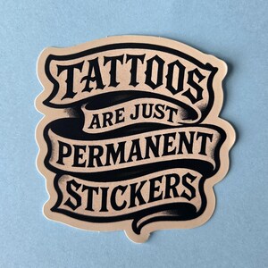 May include: A die-cut sticker featuring a beige background with black text arranged on three flowing ribbons. The text reads TATTOOS ARE JUST PERMANENT STICKERS in a bold, stylized font. The design has a smooth, matte finish with clean edges.