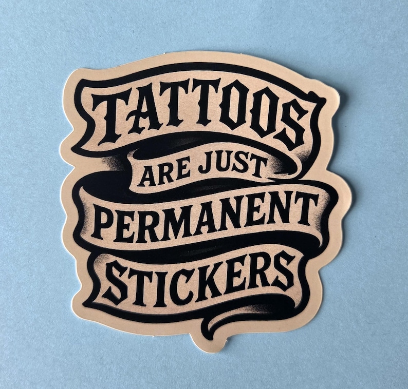 May include: A die-cut sticker featuring a beige background with black text arranged on three flowing ribbons. The text reads TATTOOS ARE JUST PERMANENT STICKERS in a bold, stylized font. The design has a smooth, matte finish with clean edges.