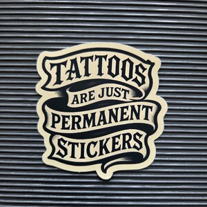May include: A die-cut sticker with a cream-colored background and black lettering arranged in a ribbon-like design. The text reads TATTOOS ARE JUST PERMANENT STICKERS in a bold, stylized font with shading that creates a layered, dimensional appearance.