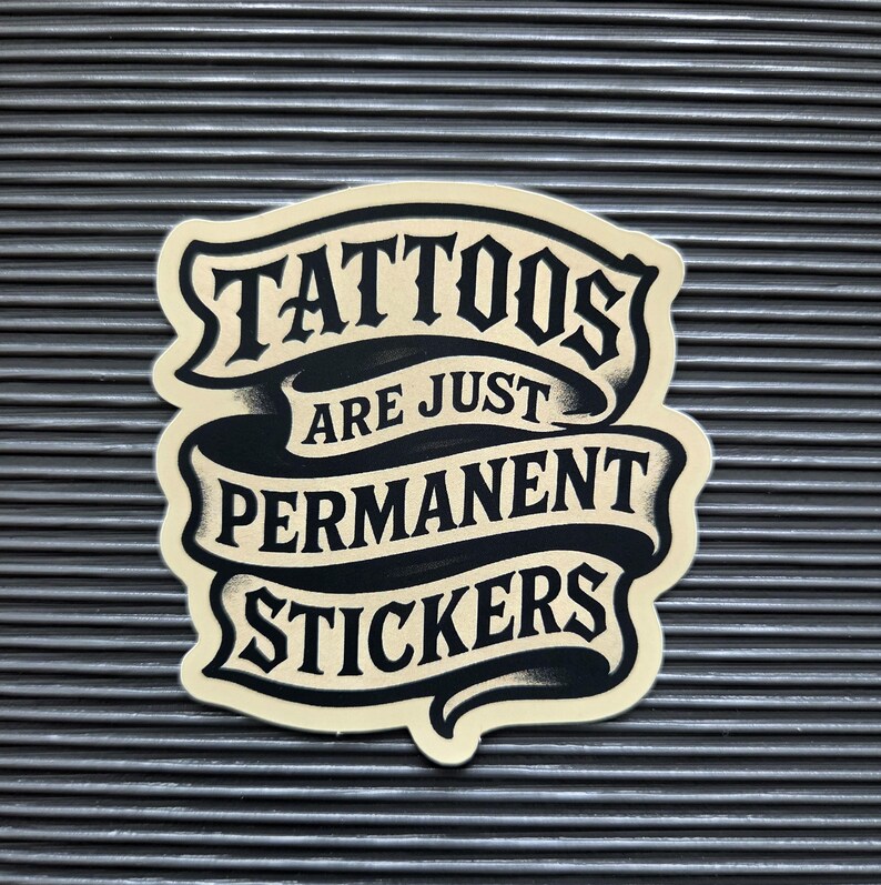 May include: A die-cut sticker with a cream-colored background and black lettering arranged in a ribbon-like design. The text reads TATTOOS ARE JUST PERMANENT STICKERS in a bold, stylized font with shading that creates a layered, dimensional appearance.
