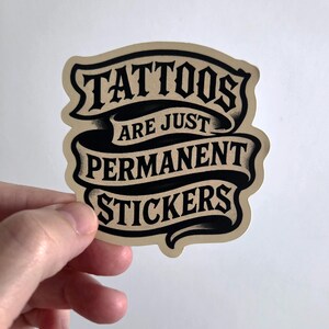 May include: A die-cut sticker with a beige background and black text arranged in three ribbon-style banners. The text reads TATTOOS ARE JUST PERMANENT STICKERS in a bold, stylized serif font with subtle black shading and a vintage-inspired aesthetic.