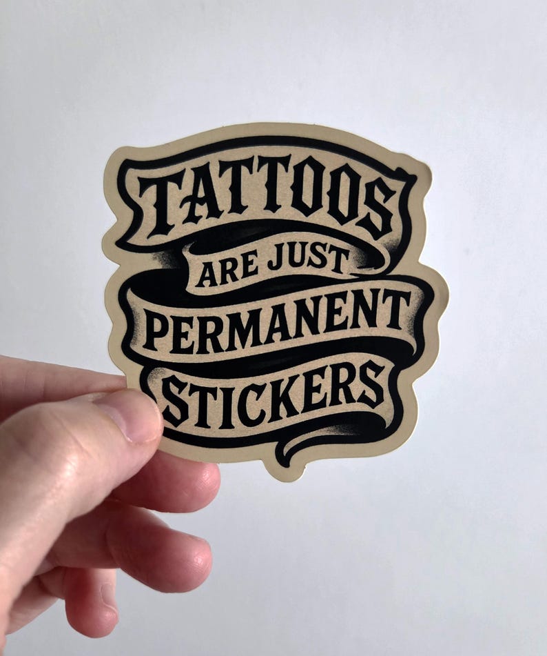 May include: A die-cut sticker with a beige background and black text arranged in three ribbon-style banners. The text reads TATTOOS ARE JUST PERMANENT STICKERS in a bold, stylized serif font with subtle black shading and a vintage-inspired aesthetic.