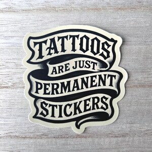 May include: A die-cut sticker featuring a black and cream-colored ribbon design with the text "TATTOOS ARE JUST PERMANENT STICKERS" printed in a bold, stylized serif font. The graphic has a distressed, ink-like texture with shading.