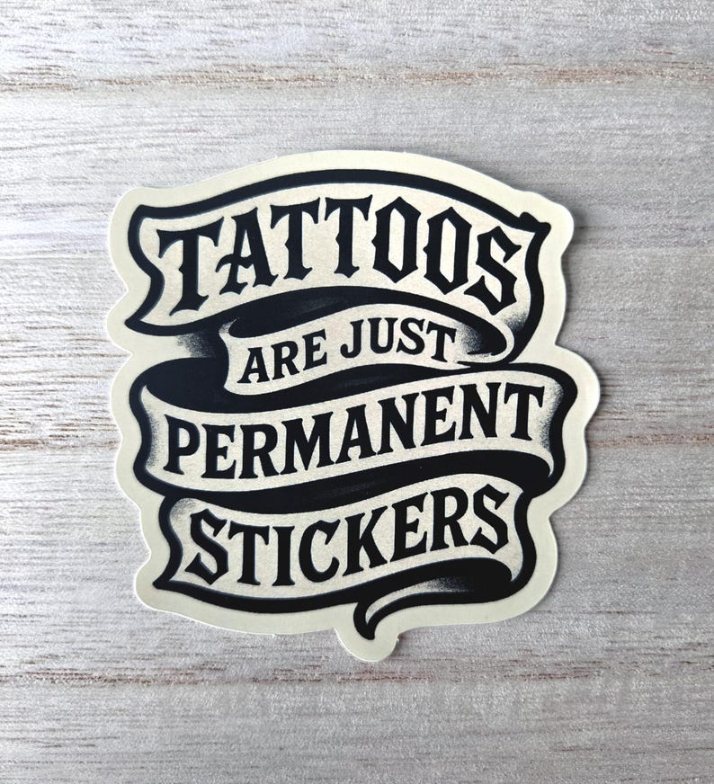 May include: A die-cut sticker featuring a black and cream-colored ribbon design with the text "TATTOOS ARE JUST PERMANENT STICKERS" printed in a bold, stylized serif font. The graphic has a distressed, ink-like texture with shading.