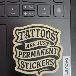 May include: A die-cut sticker featuring a beige and black banner design with the text "TATTOOS ARE JUST PERMANENT STICKERS" in a bold, stylized font. The sticker has a matte finish and is applied to the dark gray surface of a laptop keyboard.