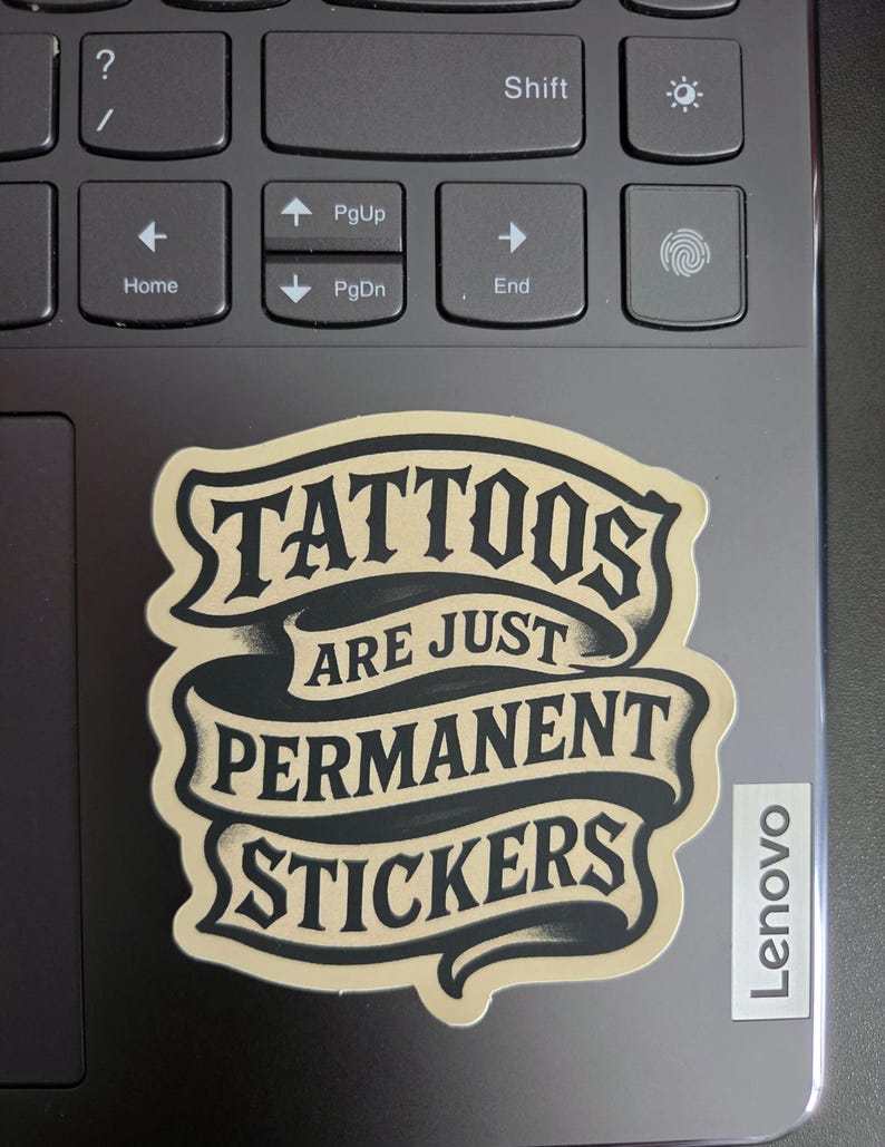 May include: A die-cut sticker featuring a beige and black banner design with the text "TATTOOS ARE JUST PERMANENT STICKERS" in a bold, stylized font. The sticker has a matte finish and is applied to the dark gray surface of a laptop keyboard.