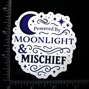 May include: A white sticker with a scalloped edge, featuring the text "Powered By Moonlight & Mischief" in navy blue. The design includes a crescent moon, stars, and decorative swirls. The sticker is on a black background.