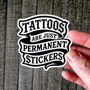 May include: A die-cut vinyl sticker featuring black lettering on a white background. The text reads TATTOOS ARE JUST PERMANENT STICKERS, arranged in three lines within a stylized ribbon banner design with black shading and bold typography.