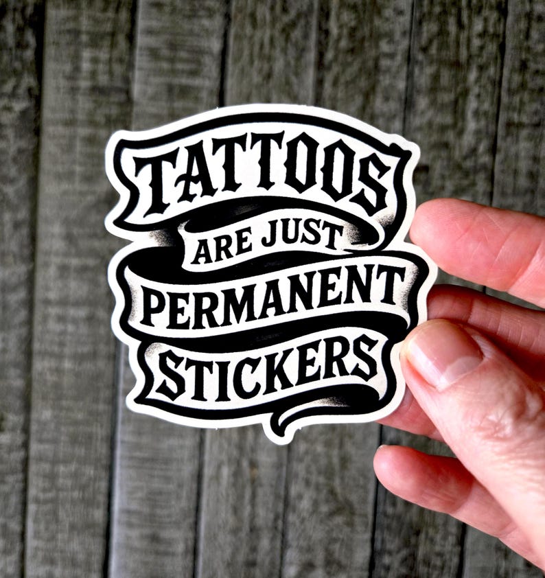 May include: A die-cut vinyl sticker featuring black lettering on a white background. The text reads TATTOOS ARE JUST PERMANENT STICKERS, arranged in three lines within a stylized ribbon banner design with black shading and bold typography.
