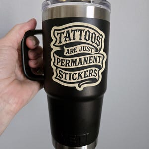 May include: A black stainless steel tumbler with a handle and a clear lid. The front features a cream-colored graphic with the text "TATTOOS ARE JUST PERMANENT STICKERS" in a stylized font. The YETI brand logo is embossed at the bottom of the cup.