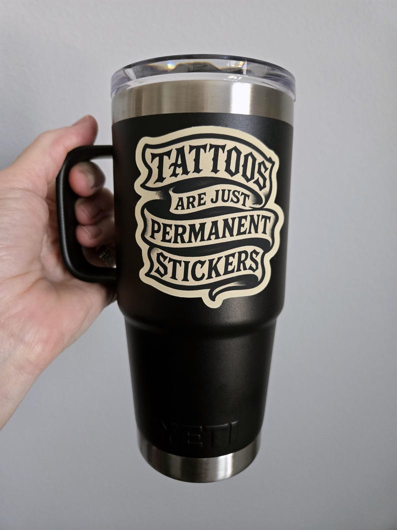 May include: A black stainless steel tumbler with a handle and a clear lid. The front features a cream-colored graphic with the text "TATTOOS ARE JUST PERMANENT STICKERS" in a stylized font. The YETI brand logo is embossed at the bottom of the cup.